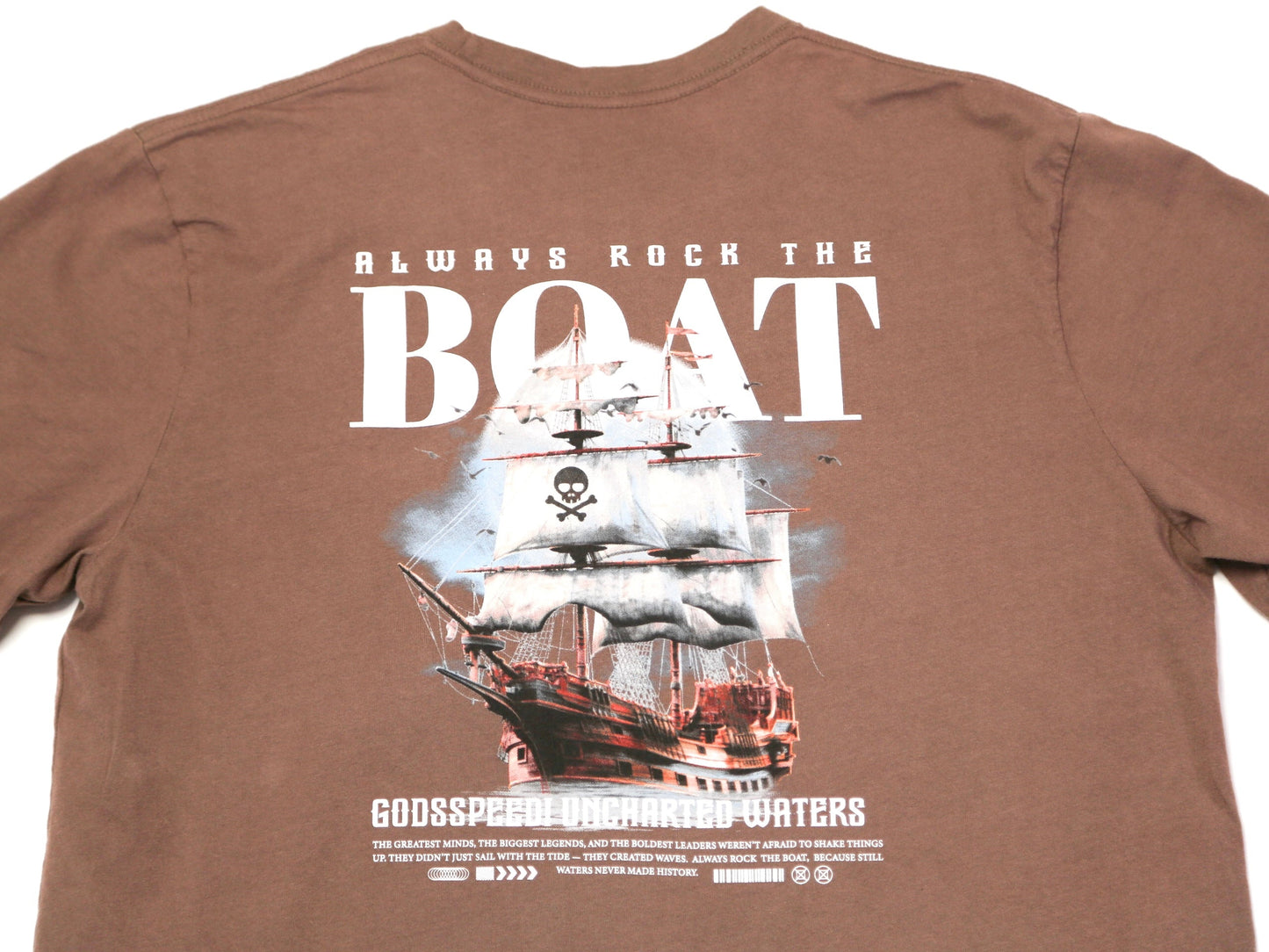 A brown short-sleeve graphic t-shirt featuring a large print of a pirate ship with white sails and the text 'ALWAYS ROCK THE BOAT' and 'GODSPEND OUR ACCUMULATED WATERS'.