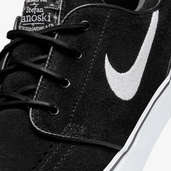 Close-up of a black suede sneaker with a large white Nike Swoosh on the side, black laces, and a visible Stefan Janoski skateboarding label on the tongue. Nike SB Janoski Black White