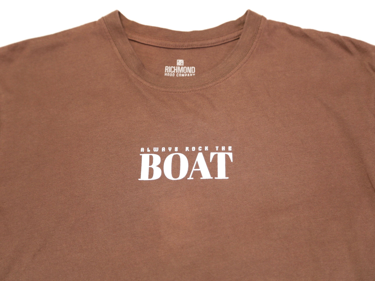 A close-up of a brown short-sleeve t-shirt with the phrase 'ALWAYS ROCK THE BOAT' printed in white across the chest. The inner neck label shows the brand 'Richmond Shirt Company'.