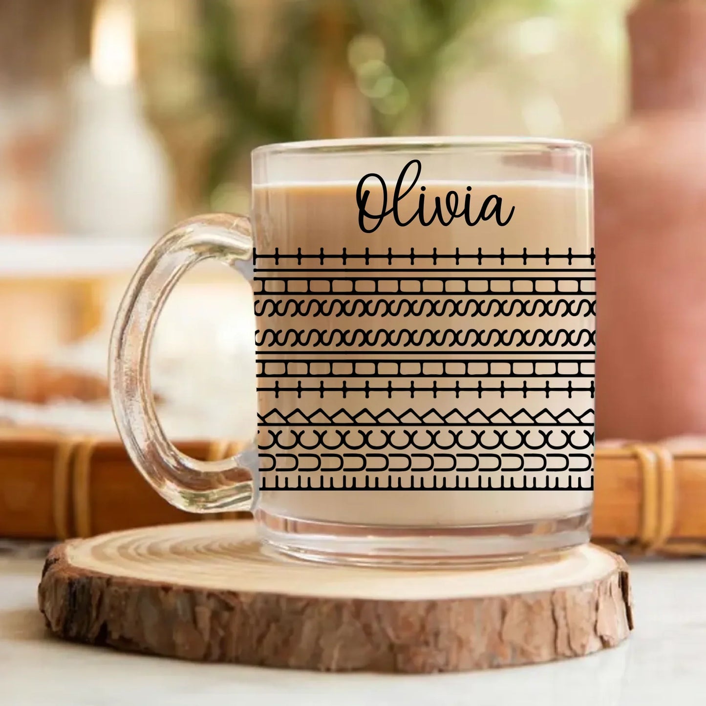 Clear mug with black pattern and 'Olivia' text, filled with a hot beverage, on a wooden coaster.