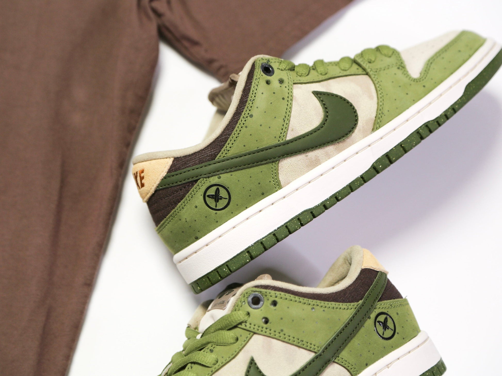 Green and beige Nike Dunk Low sneakers with dark brown accents, shown in a styled flat-lay composition with a brown fabric prop on a white background.