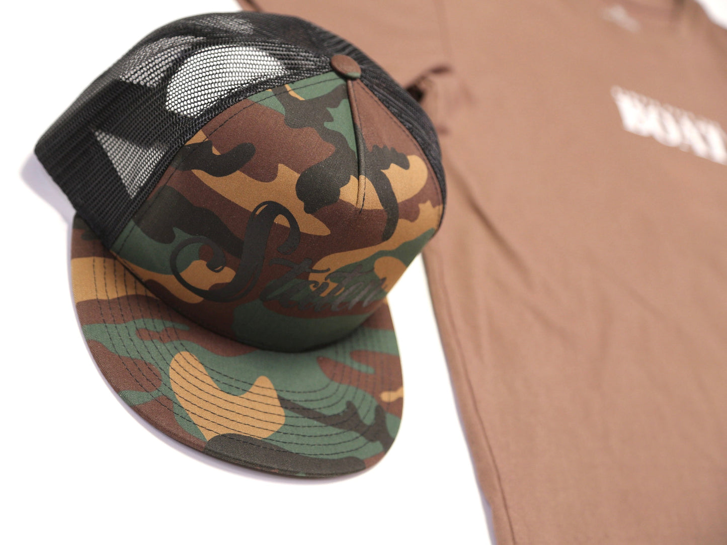 A camouflage trucker hat with a black mesh back, styled next to a brown t-shirt on a white background.