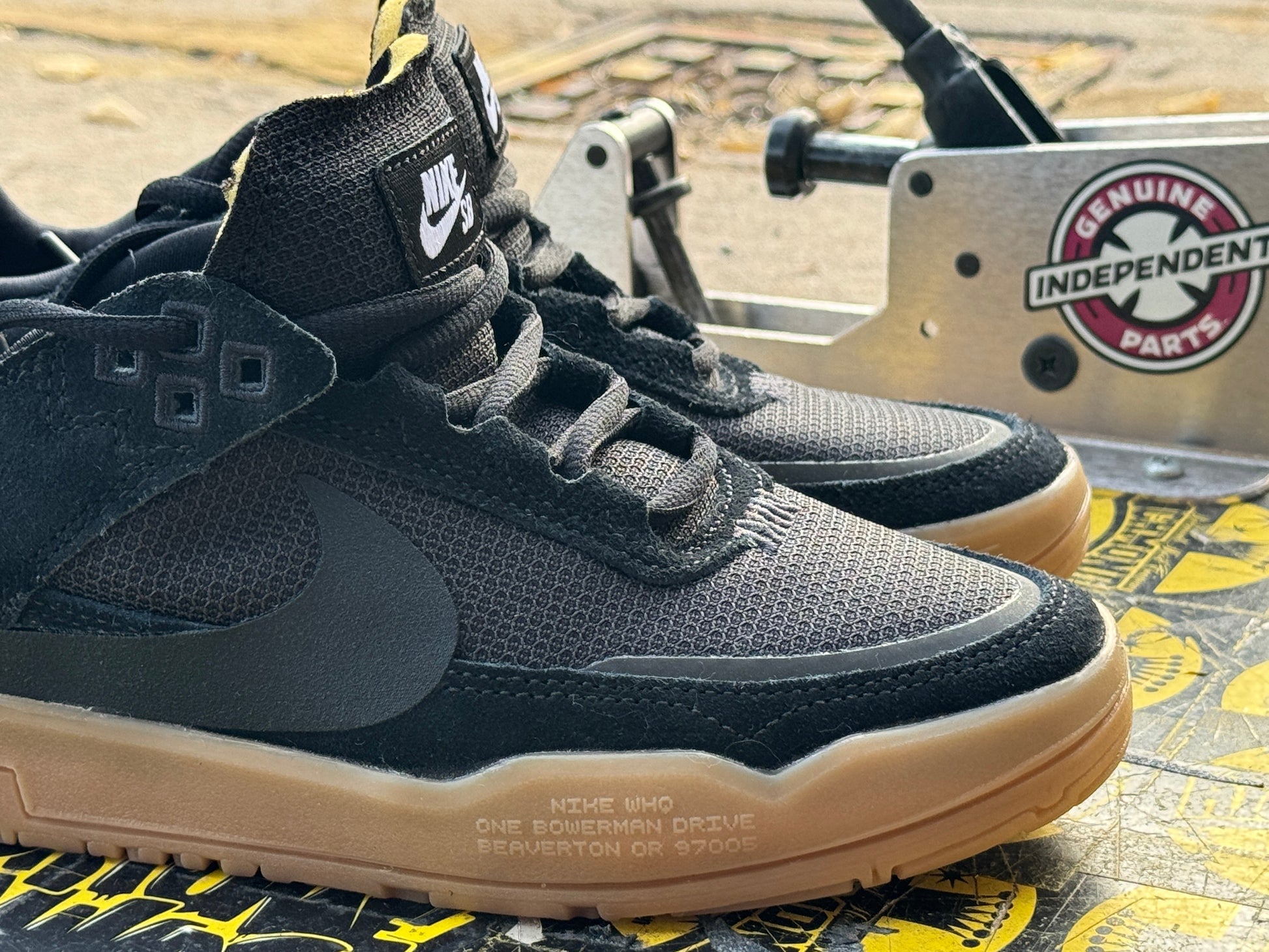 A pair of black Nike high-top sneakers with gum soles, displayed in front of a metal machine with a visible Genuine Parts logo. Nike SB Day One - Youth - FN4210-001
