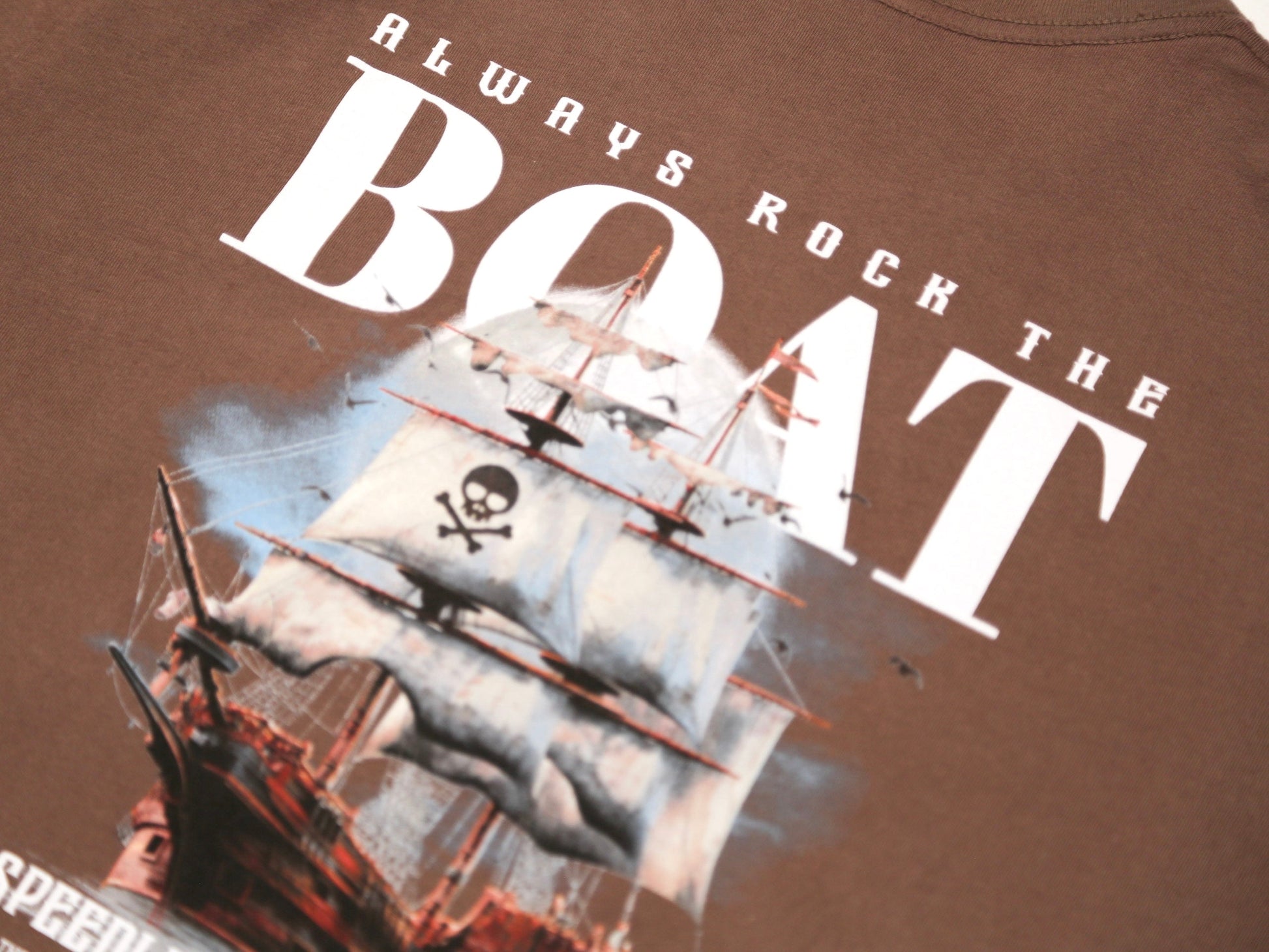 Close-up of a brown t-shirt featuring a graphic of a pirate ship with a skull and crossbones flag and the text 'ALWAYS ROCK THE BOAT' in white.