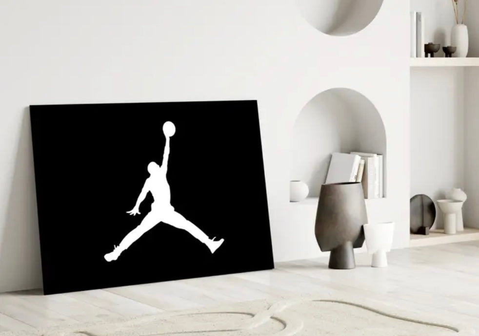 Black canvas with white silhouette of a person jumping and holding a basketball, placed on a white surface. Michael Jordan Glass Wall Art Piece for home, bedroom, bathroom decorative collectors item