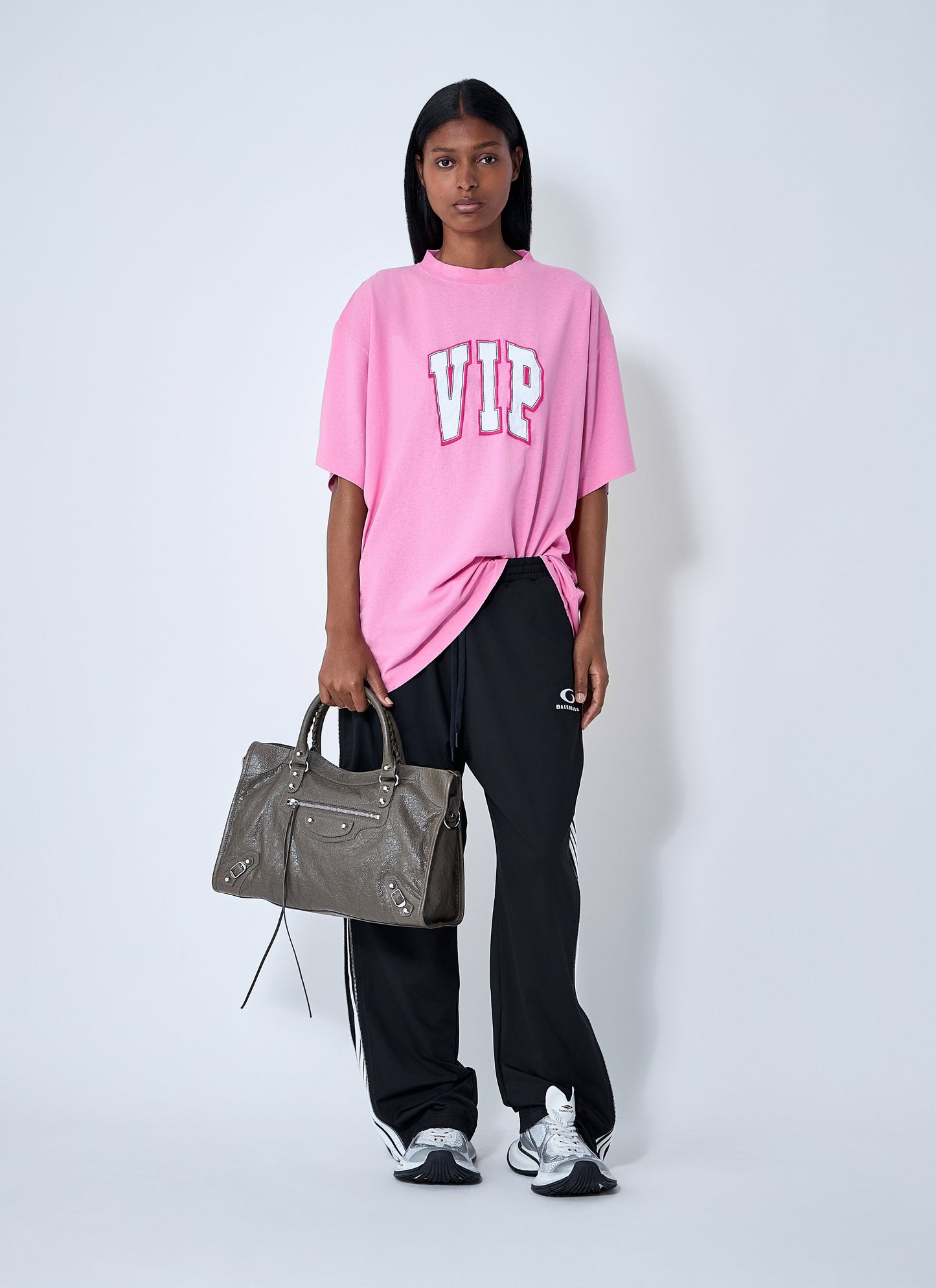 Person wearing a pink 'VIP' t-shirt and black pants, holding a gray handbag on a white background