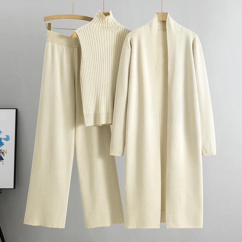 Beige clothing set with a sweater, pants, and cardigan on hangers against a gray background.