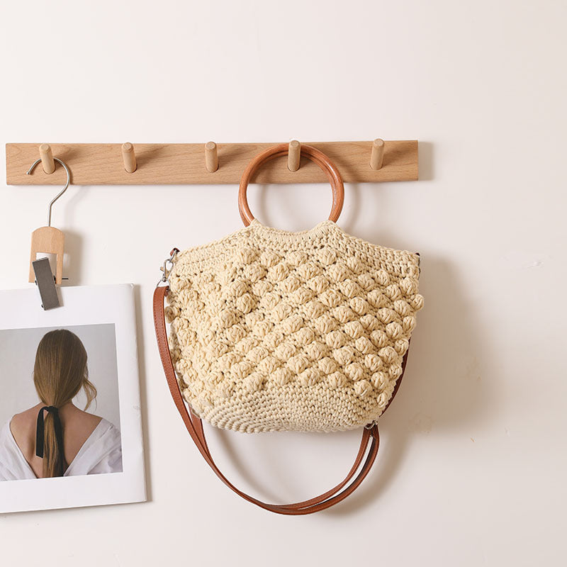 Beige woven handbag with brown handles hanging on a wooden hook against a white wall.