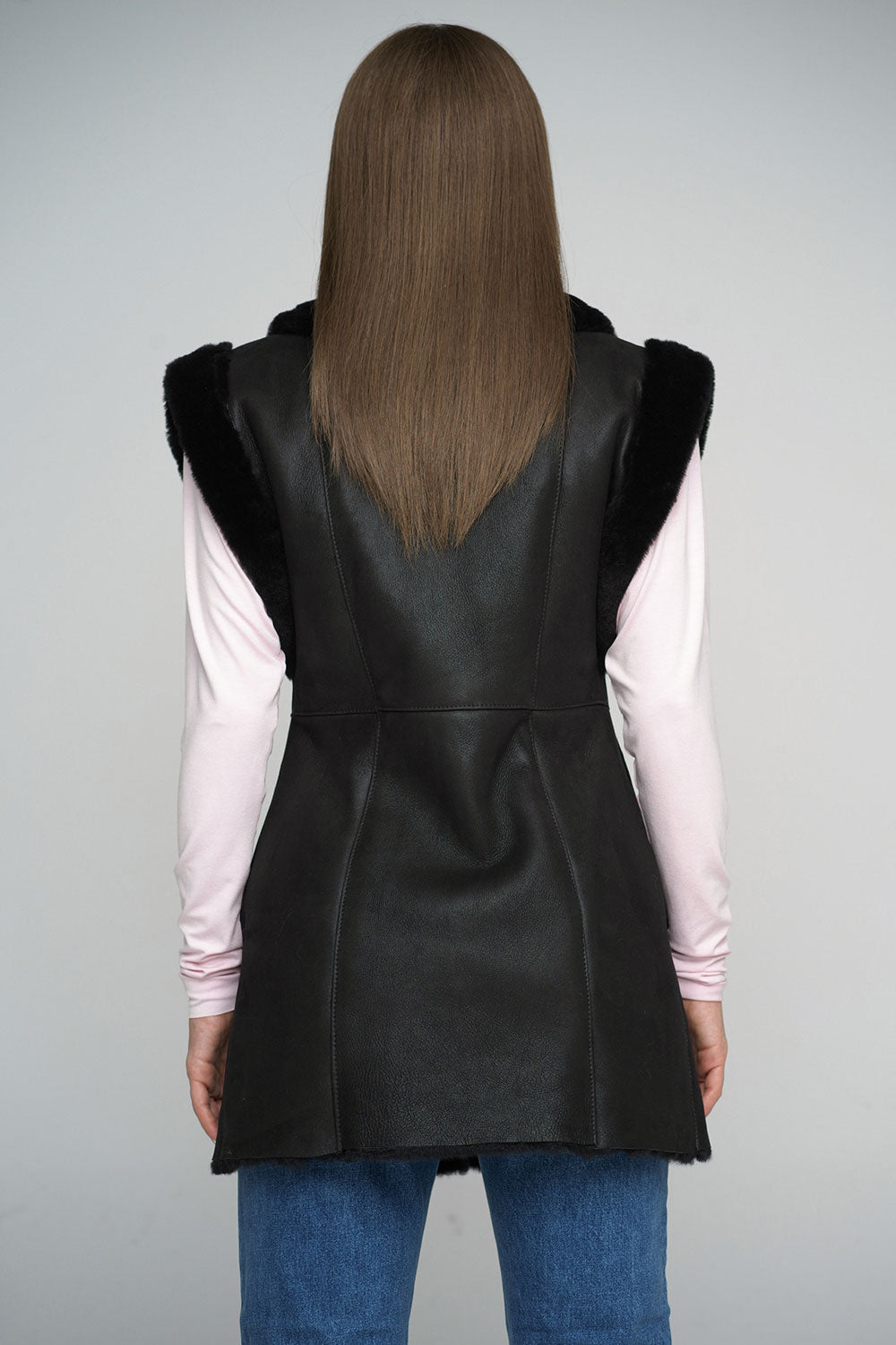 Person wearing a black leather vest over a light pink shirt and blue jeans on a gray background