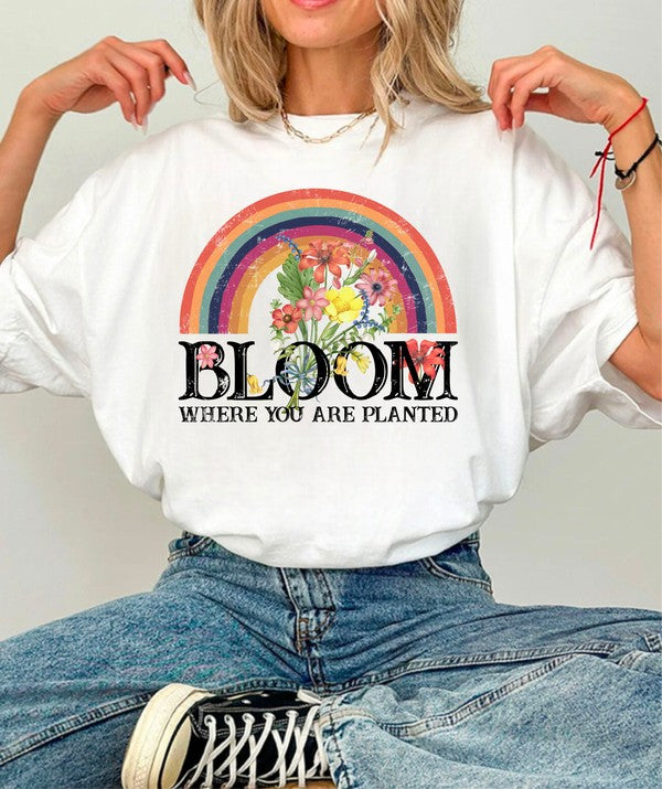 Woman wearing a white oversized graphic T-shirt featuring a colorful rainbow with blooming flowers above the inspirational phrase "Bloom Where You Are Planted." Casual faith-inspired short-sleeve tee styled with blue jeans, perfect for everyday wear, Christian fashion, spring and summer outfits, and uplifting gifts.