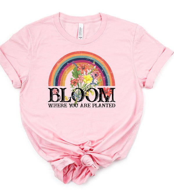 Light pink women's graphic T-shirt featuring a colorful rainbow filled with blooming flowers above the inspirational phrase "Bloom Where You Are Planted." Soft short-sleeve crew neck tee with rolled sleeves and a knotted front, displayed on a white background. Perfect for casual everyday wear, Christian faith-inspired fashion, spring and summer outfits, and uplifting gifts.