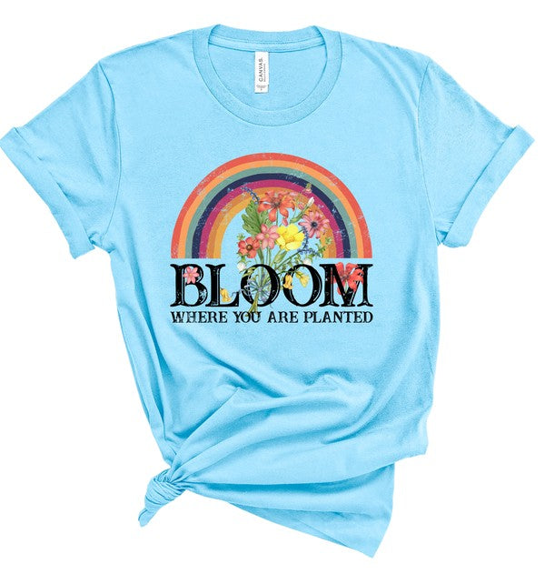 Light blue women's graphic T-shirt featuring a colorful rainbow filled with vibrant flowers above the inspirational phrase "Bloom Where You Are Planted." Soft short-sleeve crew neck tee with rolled sleeves and a knotted front, displayed on a white background. Inspirational floral faith-inspired shirt perfect for casual everyday wear, spring and summer outfits, Christian gifts, and motivational fashion.