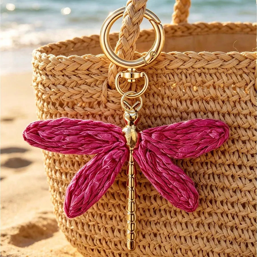 Pink dragonfly alloy bag charm with gold keyring attached to a woven straw beach tote, photographed on a sandy beach. Bohemian handbag accessory in vibrant pink with textured wings, ideal for purses, totes, backpacks, and keychains.