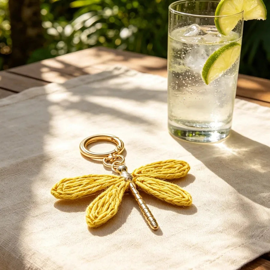 Yellow dragonfly bag charm with embroidered wings and a gold-tone alloy keyring displayed on a linen-covered table beside a refreshing glass of sparkling water with lime slices, showcasing a stylish bohemian handbag accessory.