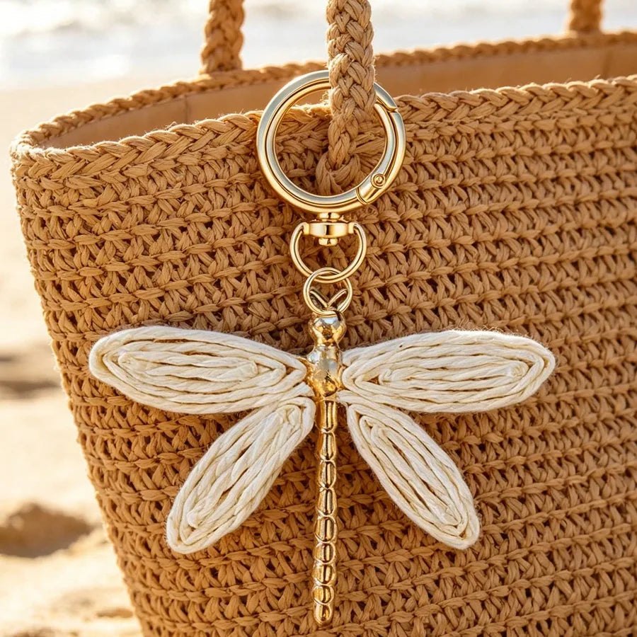 Dragonfly-shaped bohemian bag charm with woven cream wings, gold-tone alloy body, and keyring attached to a natural woven straw beach tote, creating a stylish purse accessory for handbags, backpacks, and tote bags.