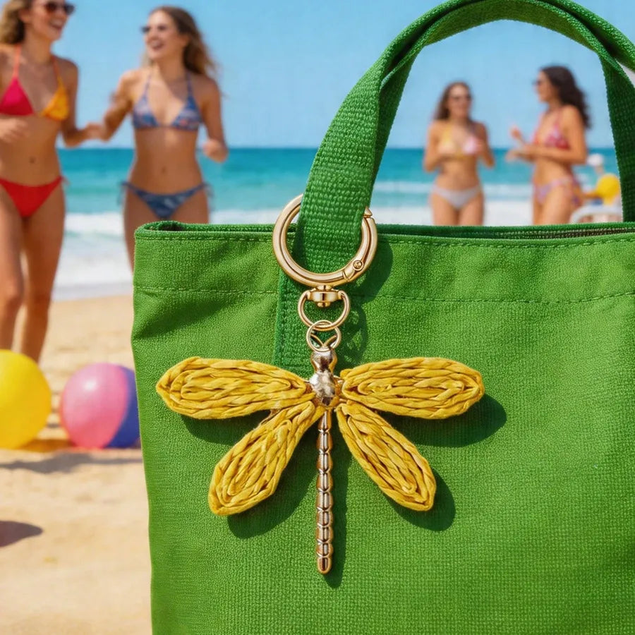 Gold dragonfly alloy bag charm attached to a vibrant green tote bag, photographed on a sunny beach with colorful balloons and people in the background. Bohemian-inspired handbag accessory, decorative purse charm, and stylish keychain pendant for totes, handbags, backpacks, and everyday fashion.