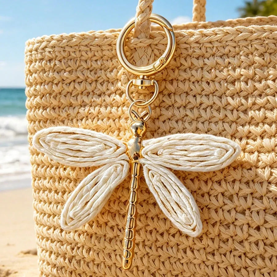 Dragonfly bag charm with woven cream wings and a gold-tone alloy body attached to a natural straw beach tote, photographed against a sunny seaside background. Bohemian purse accessory, keychain pendant, and handbag charm for women.