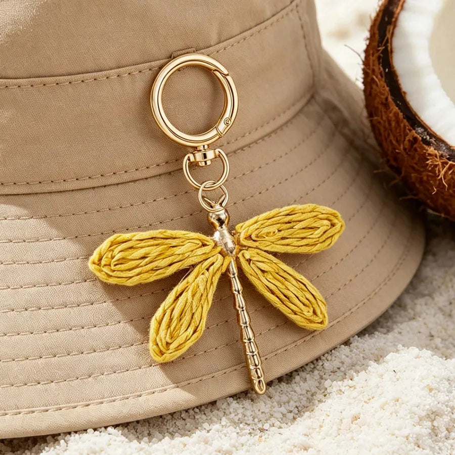 Golden yellow dragonfly alloy bag charm with a gold keyring clasp displayed on a beige bucket hat, surrounded by white sand and a coconut shell, showcasing a bohemian-inspired handbag and keychain accessory.