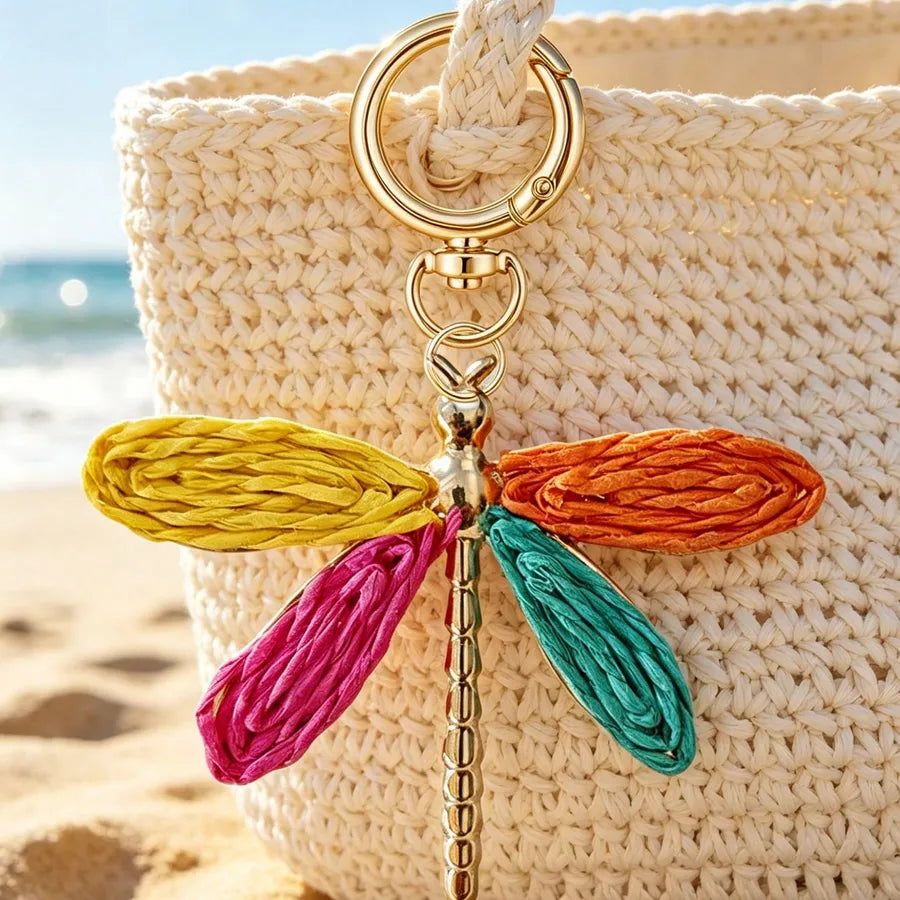 Colorful dragonfly bag charm with a gold-tone alloy keyring clipped to a woven straw beach tote, featuring vibrant woven wings in yellow, orange, teal, and magenta for a bohemian-inspired handbag accessory.