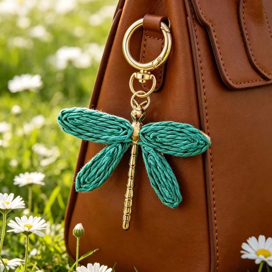 Teal woven dragonfly bag charm with gold-tone alloy hardware attached to a brown leather handbag, photographed outdoors with white flowers and lush greenery. Bohemian-style keychain accessory for purses, totes, backpacks, and handbags.