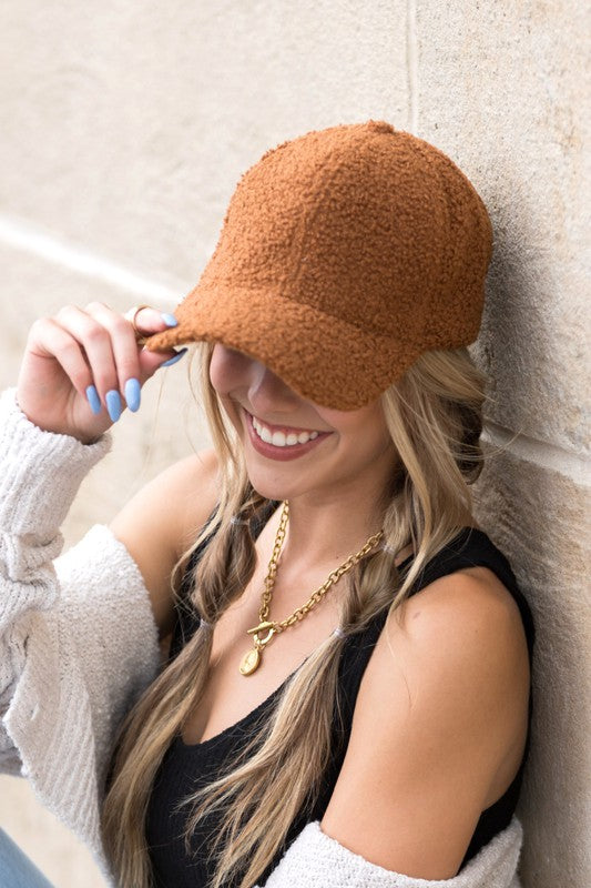 Woman wearing a dark camel boucle sherpa teddy bear knit baseball cap with braided blonde hair, smiling while leaning against a stone wall in a casual outfit.
