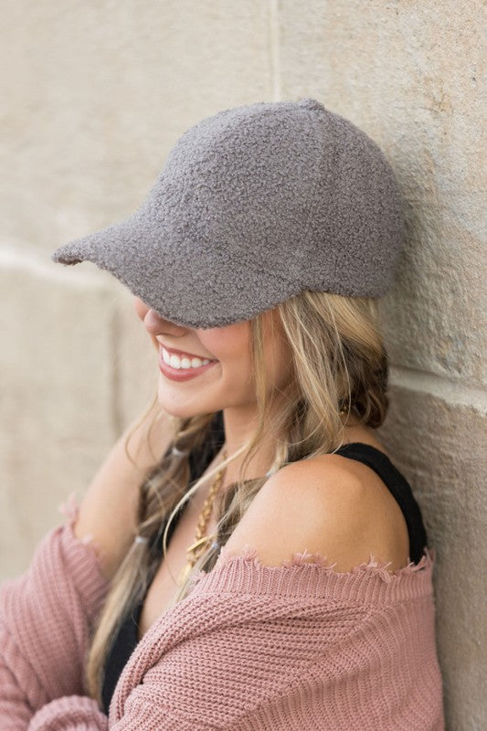 Woman wearing a gray boucle sherpa teddy bear knit baseball cap with braided blonde hair, smiling while leaning against a stone wall in a casual outfit.