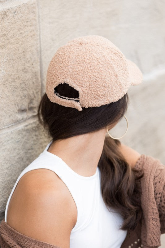 Woman wearing a beige boucle sherpa teddy bear knit baseball cap from the back with a low ponytail, white sleeveless top, and brown cardigan against a stone wall.
