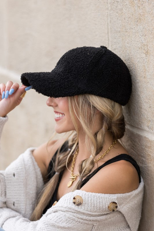 Woman wearing a black boucle sherpa teddy baseball cap with braided blonde hair, smiling while leaning against a stone wall in a casual outfit.