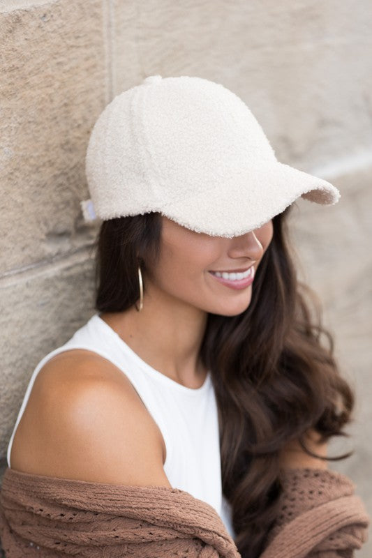 Woman wearing a cream boucle sherpa teddy knit baseball cap with a white sleeveless top and brown cardigan, smiling while leaning against a stone wall.