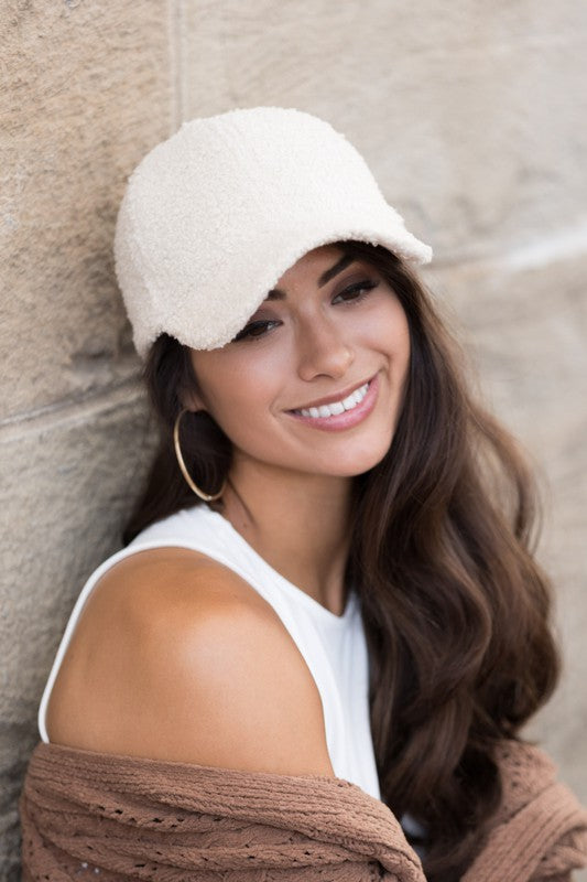 Woman wearing a cream boucle sherpa teddy bear knit baseball cap with a white sleeveless top and brown cardigan, smiling while leaning against a stone wall.