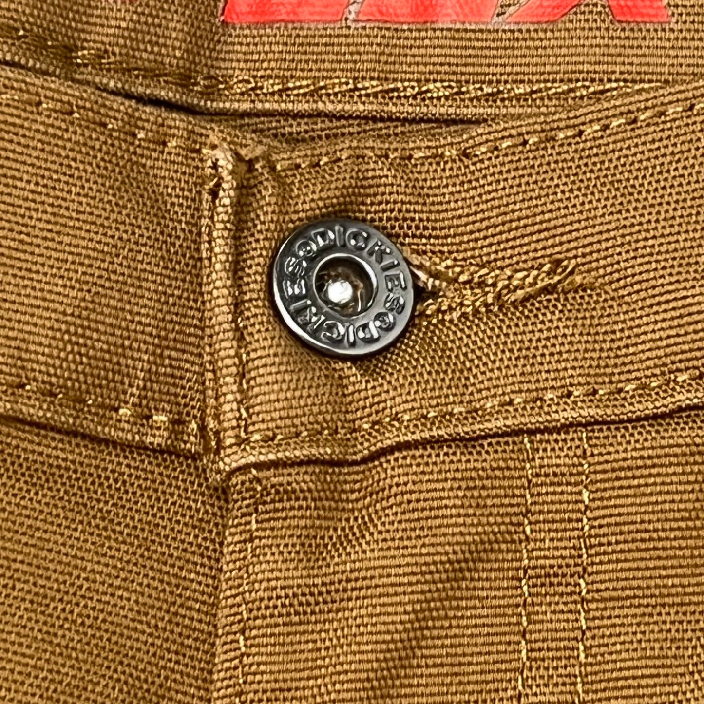 Close-up of a button on brown fabric with 'Dickies' branding. Brown pants with Flex and Dickies branding on a beige background