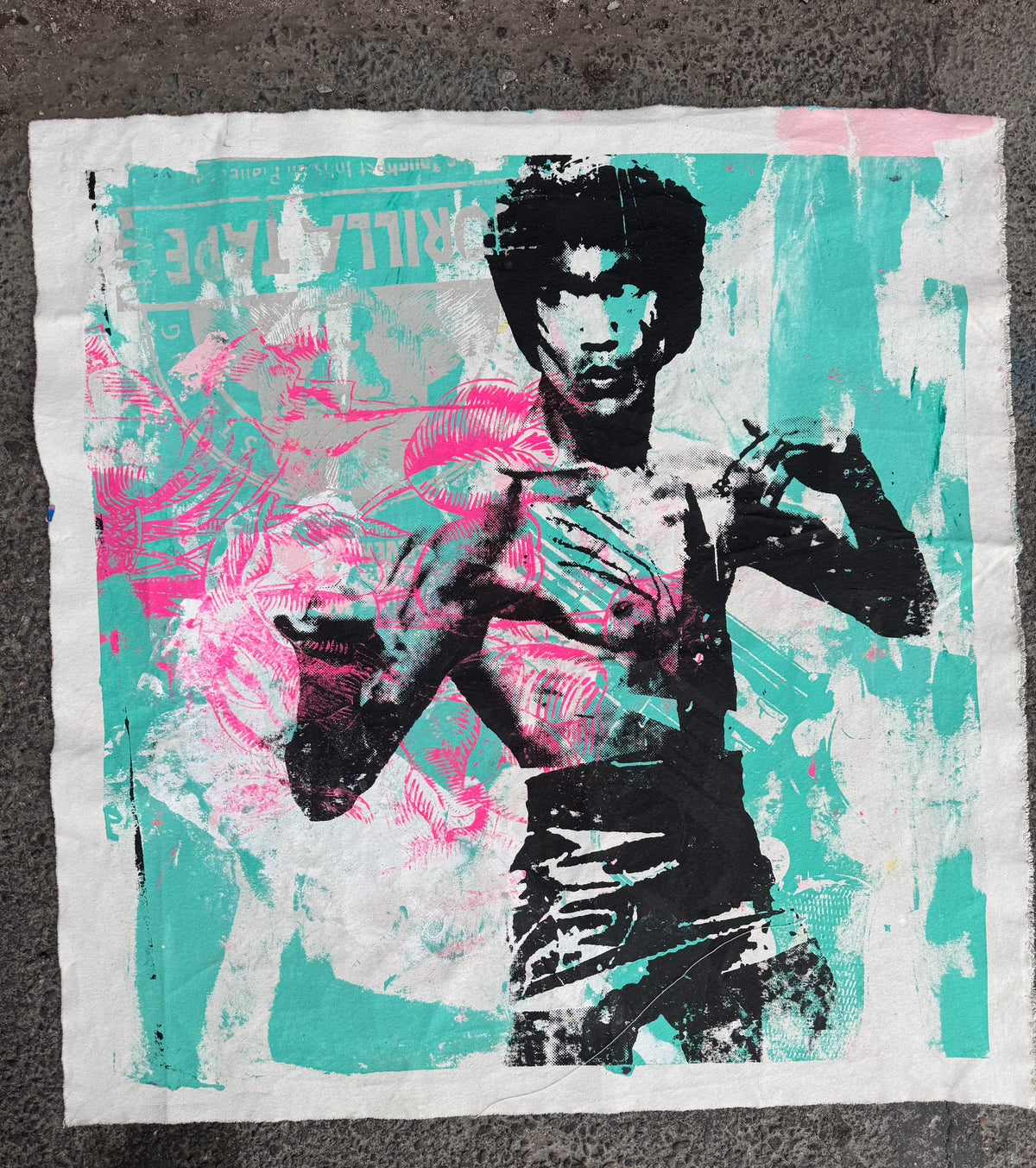 A square textile print or patch featuring a stylized pop-art portrait of Bruce Lee in black, with a turquoise background and bright pink abstract brushstrokes, laid flat on a dark textured surface. Bruce Lee & The Green Gun - Silk Screen & Spray Paint on canvas, 2025