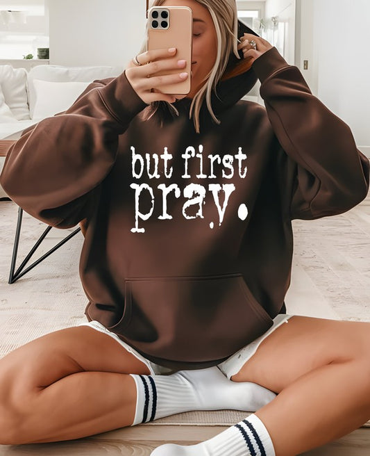Woman wearing an oversized brown hoodie with the phrase "But First Pray" in bold white lettering, sitting indoors and taking a mirror selfie. Features a cozy ring-spun cotton blend fleece pullover with a front kangaroo pocket, adjustable drawstring hood, ribbed cuffs, and a relaxed unisex fit, ideal for Christian faith-inspired casual wear.