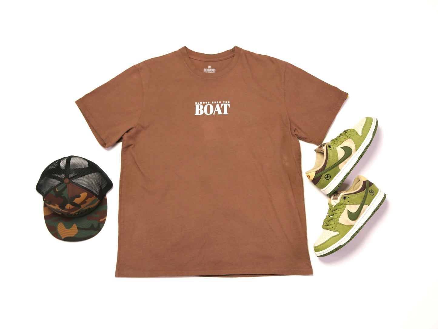 A flat-lay product presentation featuring a brown crewneck t-shirt with 'BOAT' printed in white on the chest, styled with a camouflage trucker hat and a pair of green Nike Dunk sneakers on a white background.