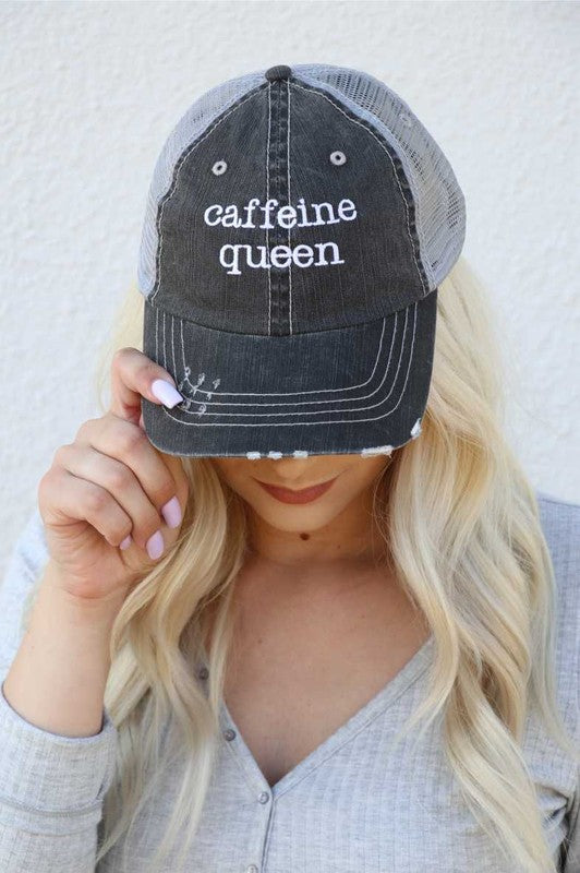 Blonde woman wearing a distressed black and gray mesh trucker hat embroidered with the words "caffeine queen," adjusting the brim while modeling the adjustable cotton and polyester cap. Caffeine Queen Trucker Hat 80% cotton/20% polyester, Herringbone fabric 100% polyester mesh back Velcro closure