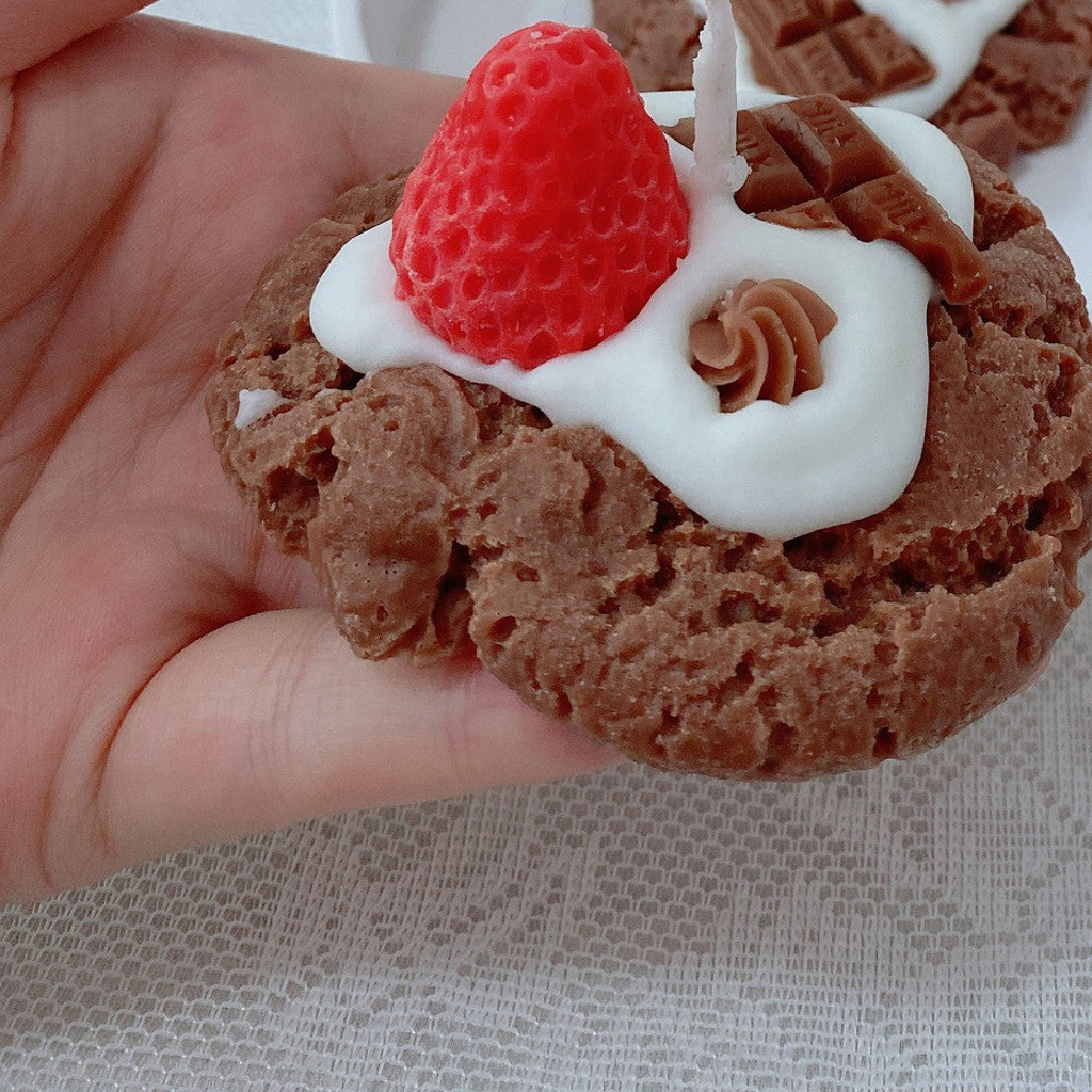 Chocolate cookie with white frosting and a strawberry on top held by a hand. Chocolate desserts with whipped cream and raspberries on a white tablecloth. Handmade Strawberry Cake Aromatic Candle Decoration.