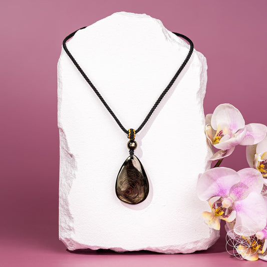 A teardrop-shaped smoky brown hypersthene gemstone pendant necklace on a black braided cord, displayed on a white stone block with pink orchids on a pink background.