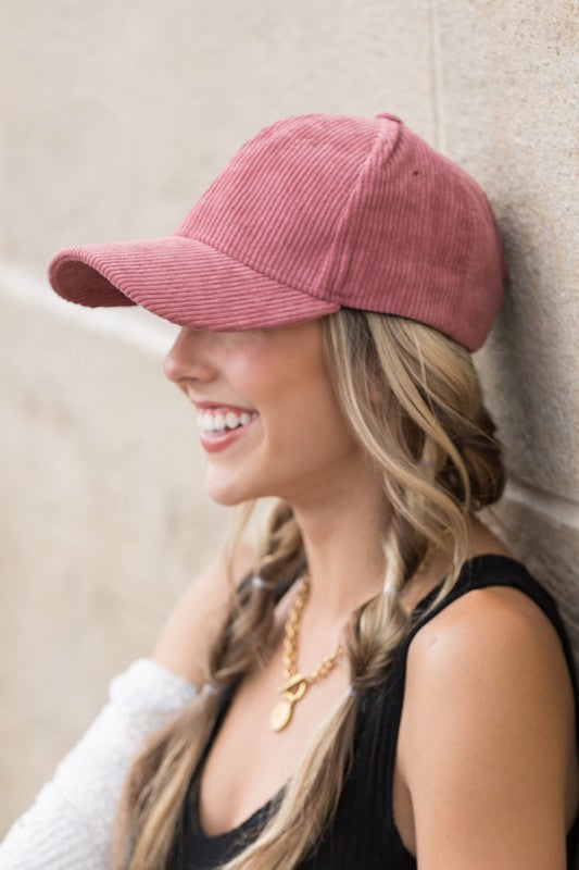 Woman smiling in a pink ribbed corduroy boyfriend baseball cap, white sleeveless top, and brown cardigan outdoors.