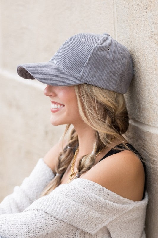 Woman wearing a grey corduroy boyfriend baseball cap with a relaxed fit, paired with a neutral sweater against a beige wall.