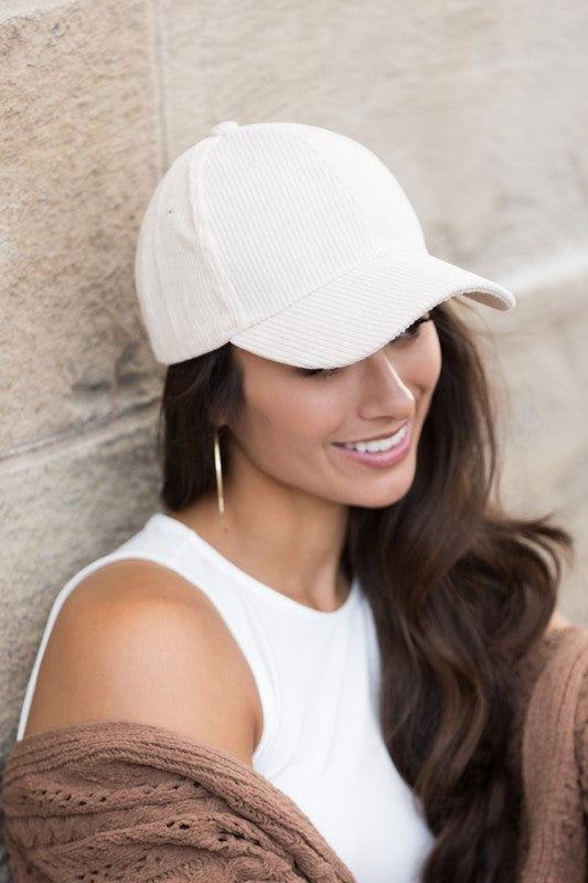 Woman wearing a white ribbed corduroy baseball cap, white sleeveless top, and brown knit cardigan, smiling while leaning against a beige stone wall.