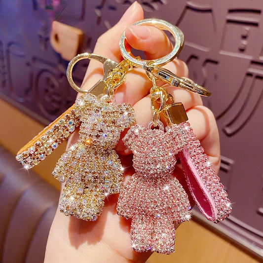 Sparkling rhinestone bear keychains in gold and pink with matching tassels, held in a hand. Decorative key ring charms for keys, handbags, backpacks, purses, or car accessories against a textured background.