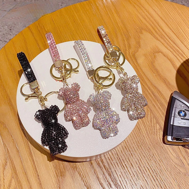 Sparkling rhinestone bear keychains with glitter wrist straps displayed on a white round tray. Four colorful bear charms in black, pink, iridescent, and champagne gold are shown beside a car key fob, ideal for keys, handbags, backpacks, purses, or as decorative bag charms.