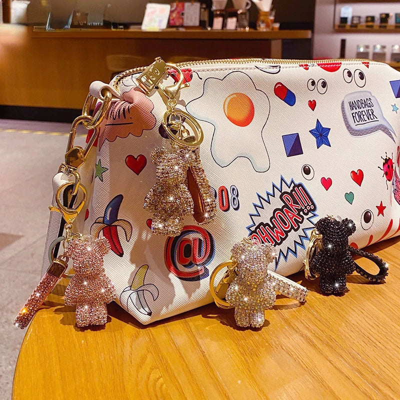 Sparkling rhinestone bear keychains with tassel accents hanging from a white cartoon-print cosmetic pouch on a wooden table, available in gold, rose gold, and black finishes.