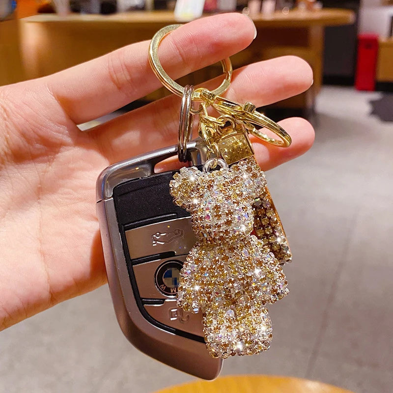 Sparkling rhinestone teddy bear keychain attached to a gold key ring and car key fob, held in a hand indoors. Decorative bear charm with tassel, ideal for handbags, backpacks, purses, or keys.