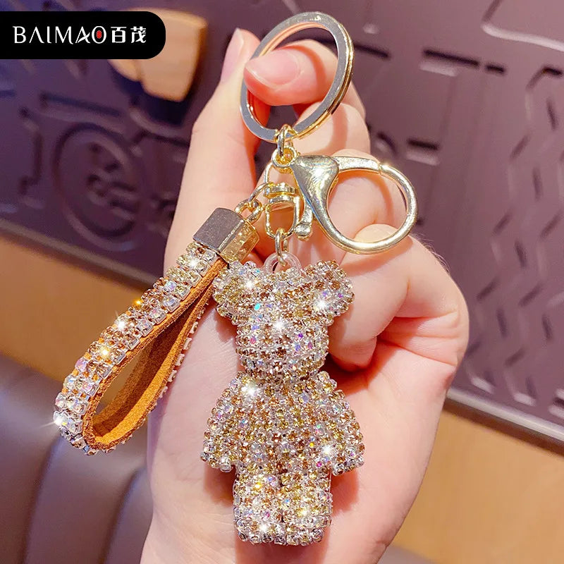 Hand holding a gold rhinestone teddy bear keychain with a sparkling tassel charm and metal key ring, attached to a handbag or car keys. Decorative bag charm with a cute bear pendant and crystal accents.