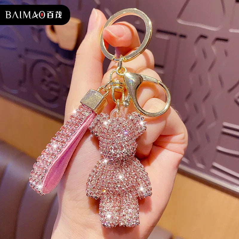Sparkling pink rhinestone teddy bear keychain with a matching rhinestone tassel, gold key ring, and lobster clasp, held in a hand against a softly blurred background. Cute decorative charm for keys, handbags, backpacks, purses, or car keys.