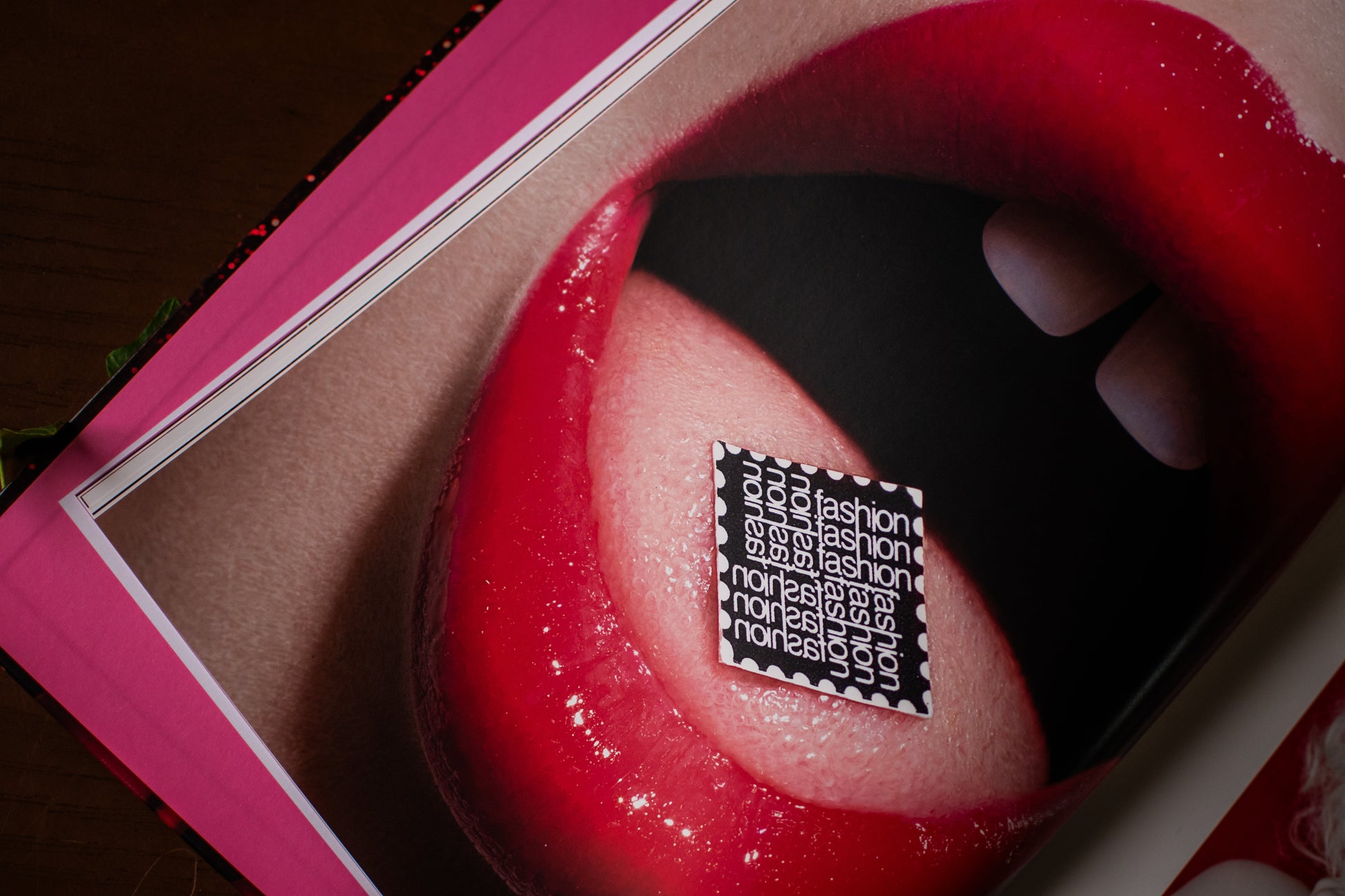 A close-up of glossy red lips with a small black-and-white patterned square sticker placed on the tongue that reads 'fashion', photographed inside a pink frame or book-like object.