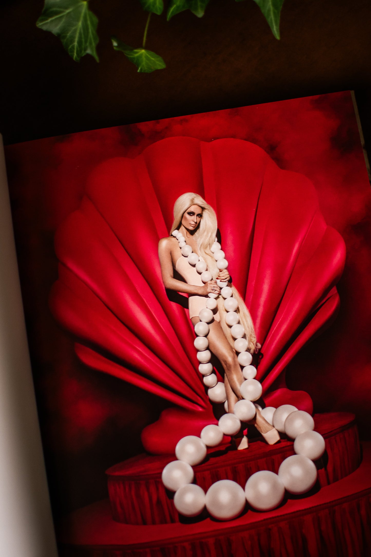 A fashion editorial-style photo of Paris Hilton featuring her modeling standing inside a large red shell-like backdrop, draped with a long white beaded necklace, with a matching beaded ring on the floor. The scene is dominated by deep red tones with white bead accents. Book title is 'High Gloss: The Art of Vijat Mohindra'