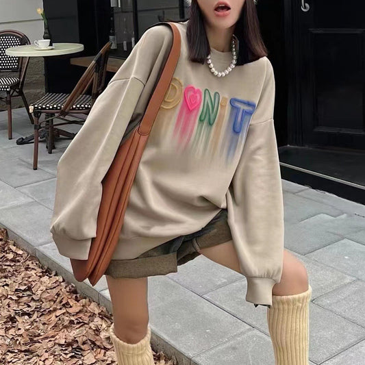 Person wearing a beige sweatshirt with colorful text, sitting outdoors on a sidewalk. Dripping Inspiration Embroidery Sweatshirt - "Somebody Love You" Graffiti theme dripping letters