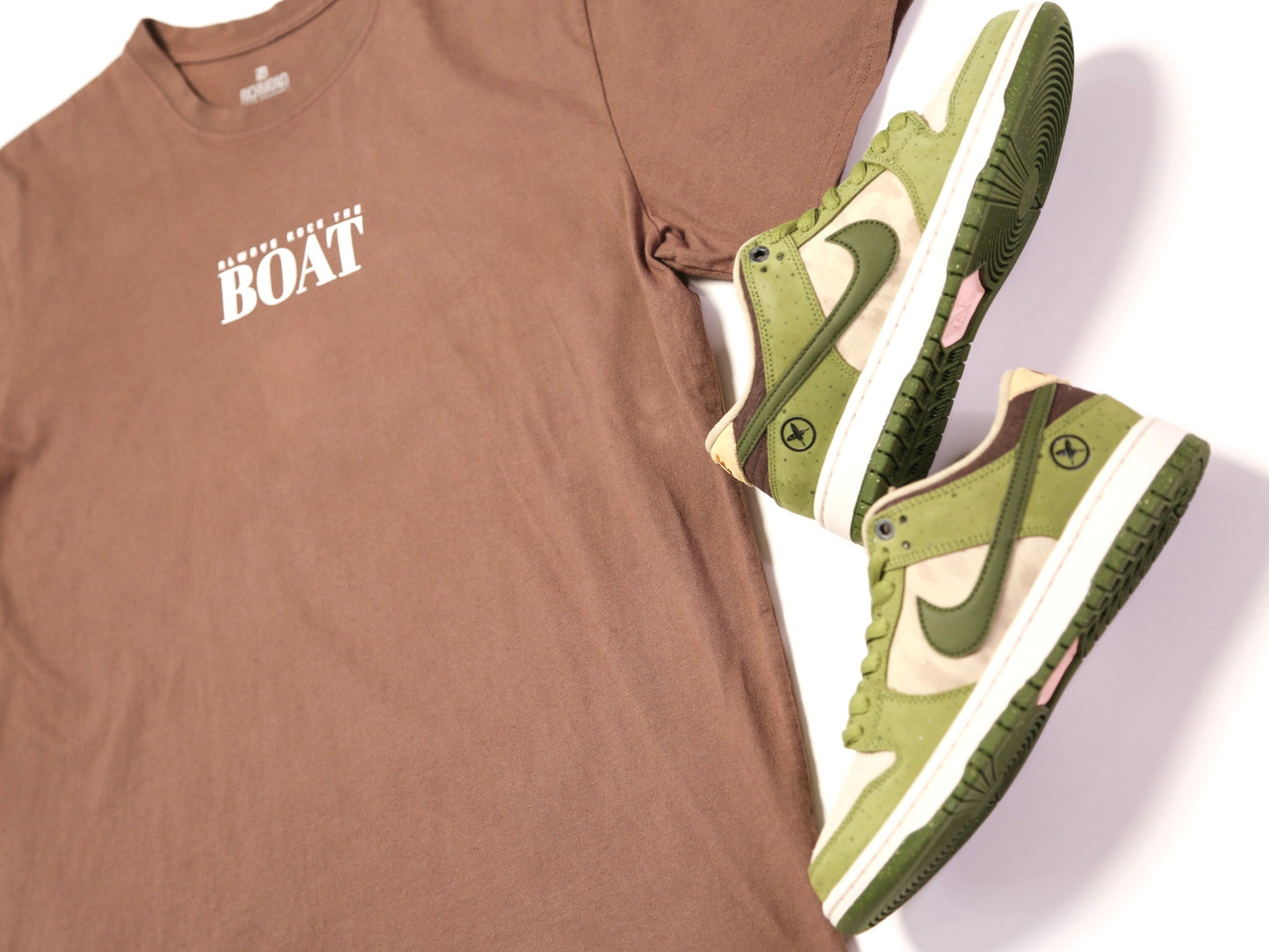 A brown crewneck t-shirt with a white 'BOAT' graphic on the chest, styled next to a pair of green and beige Nike Dunk Low x Travis Scott 'Cactus Jack' sneakers on a white background.